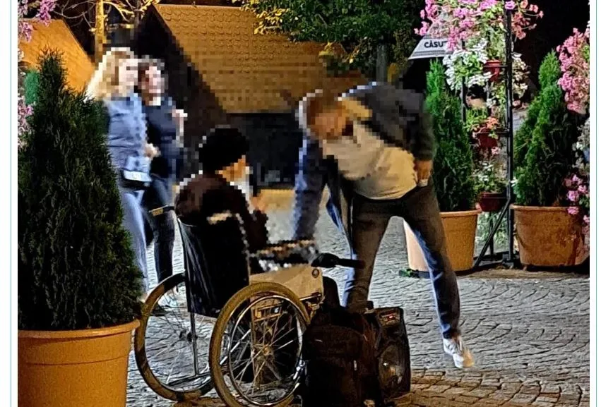 Un bărbat în pantofi albi stă aplecat lângă o persoană aflată într-un scaun cu rotile pe un pavaj, lângă câteva ghivece cu plante.