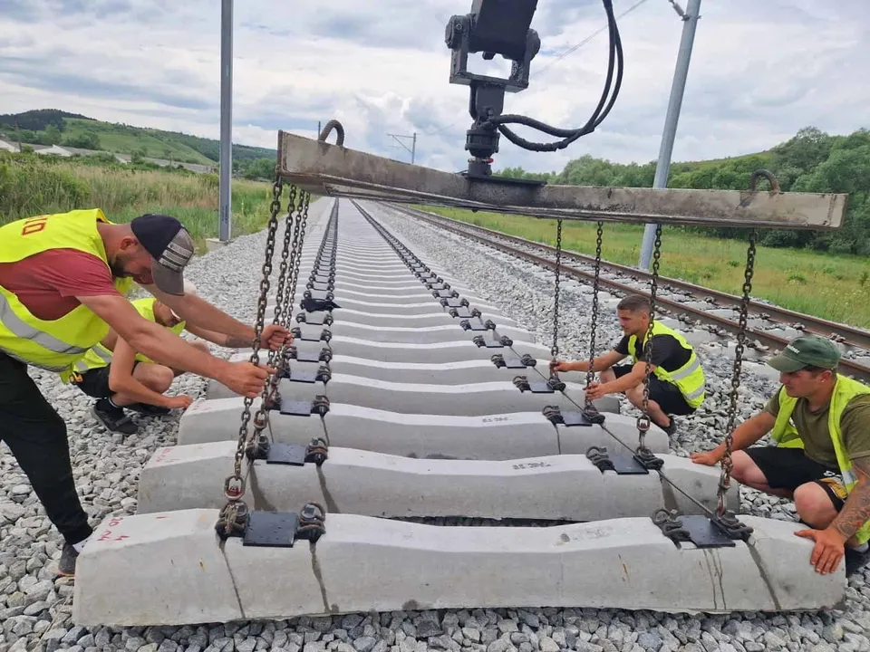 Patru bărbați cu veste reflectorizante manipulează elemente de beton suspendate prin lanțuri pe o cale ferată.