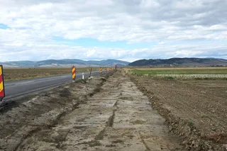 O porțiune de drum neasfaltat și noroioasă se întinde pe lângă o șosea asfaltată, sub un cer înzăpezit de nori.