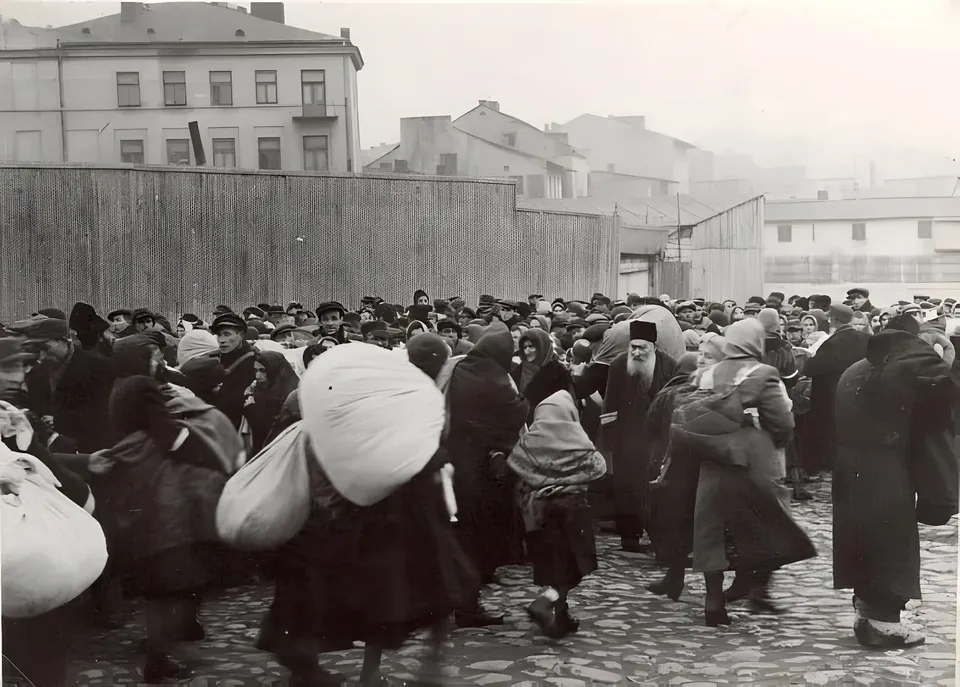 O mulțime de persoane purtând haine închise și pălării merge pe un drum pavat, purtând saci mari și pachete voluminoase.