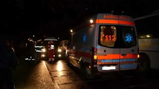 O dubă albă de ambulanță cu inscripții portocalii și numărul 112 este parcată pe un drum umed noaptea, lângă alte vehicule de serviciu.