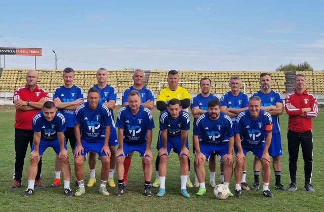 Un grup de bărbați îmbrăcați în echipamente sportive albastre și roșii pozează pe un teren de fotbal verde în fața unor tribune goale.