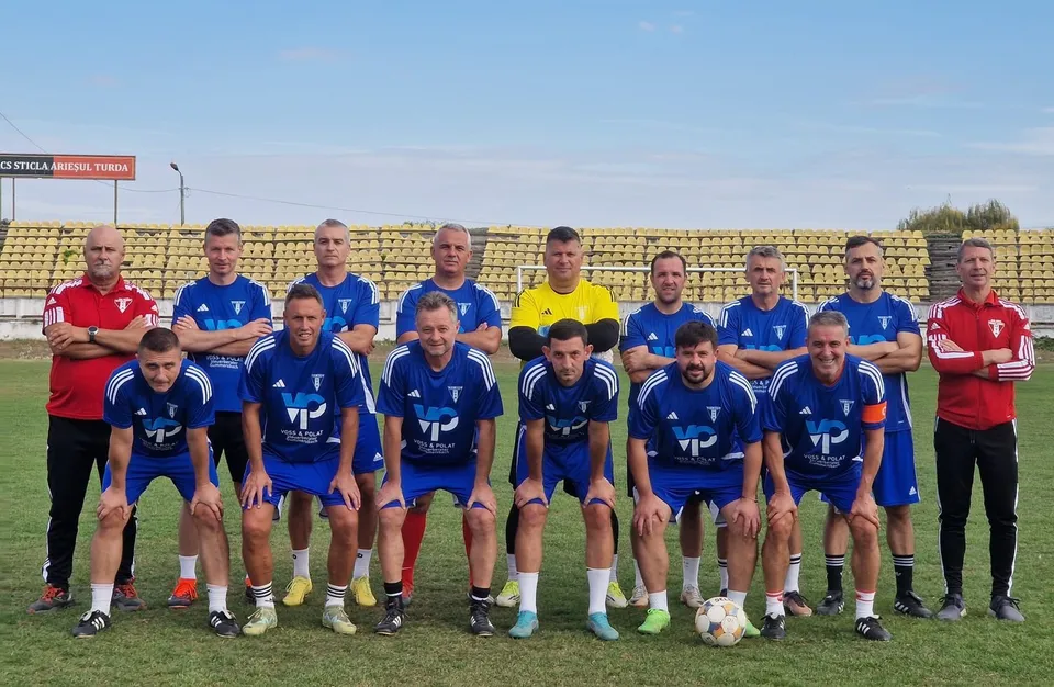 Un grup de bărbați îmbrăcați în echipamente sportive albastre și roșii pozează pe un teren de fotbal verde în fața unor tribune goale.