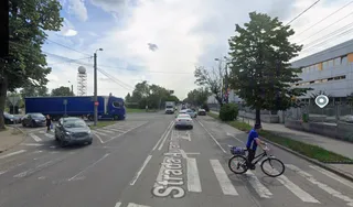 Un biciclist circulă pe o stradă asfaltată lângă un trotuar, în timp ce mașini și un camion albastru sunt parcate sau circulă în zonă.