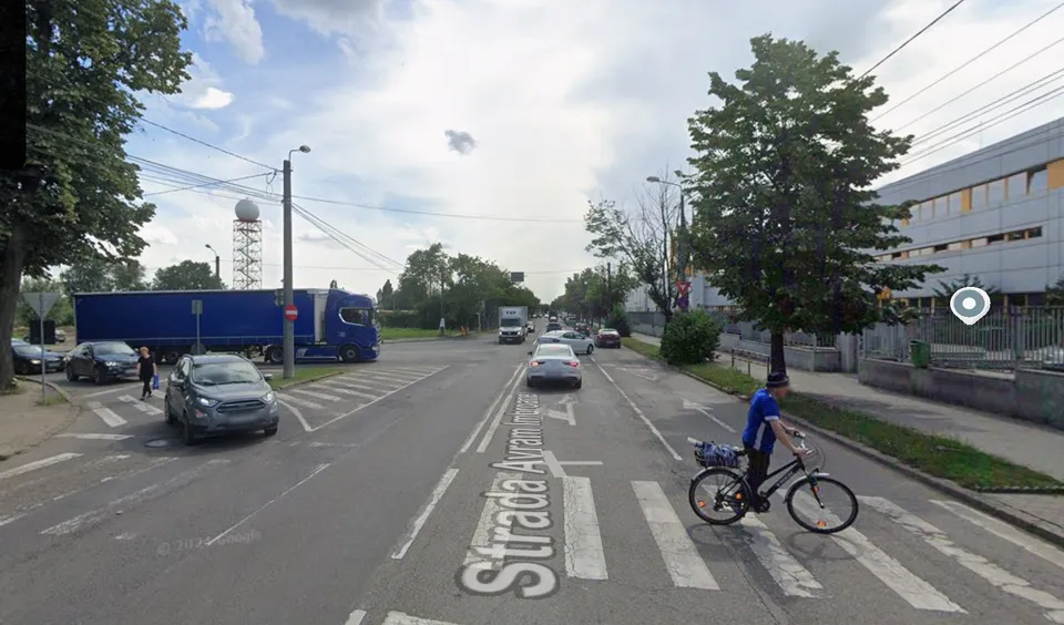 Un biciclist circulă pe o stradă asfaltată lângă un trotuar, în timp ce mașini și un camion albastru sunt parcate sau circulă în zonă.