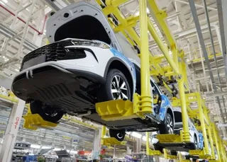 Un autoturism alb și albastru este suspendat pe o linie de asamblare industrială de culoare galbenă într-o fabrică.