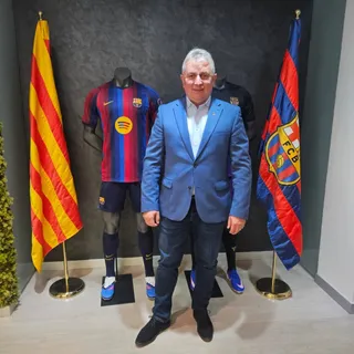 Un bărbat cu ochelari și sacou albastru stă în picioare între un manechin cu tricou de fotbal și un steag colorat.