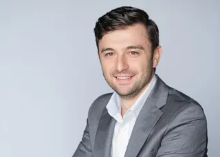 Un bărbat cu părul scurt și brunet, purtând o cămașă albă și un sacou gri, zâmbește în fața unui fundal neutru de culoare deschisă.