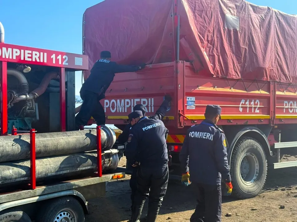 Patru persoane în uniforme închise pregătesc un camion roșu cu inscripția POMPIERII și tuburi negre de pe un trailer.