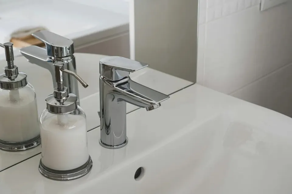 Două robinete cromate și două dozatoare de săpun cu lichid alb sunt așezate pe o chiuvetă de ceramică albă.