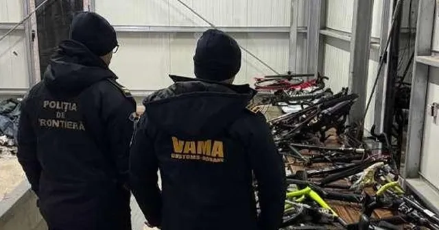 Două persoane purtând jachete negre cu inscripții galbene privesc un grup de biciclete depozitate pe o suprafață de lemn.