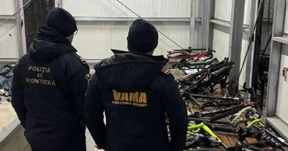 Două persoane purtând jachete negre cu inscripții galbene privesc un grup de biciclete depozitate pe o suprafață de lemn.