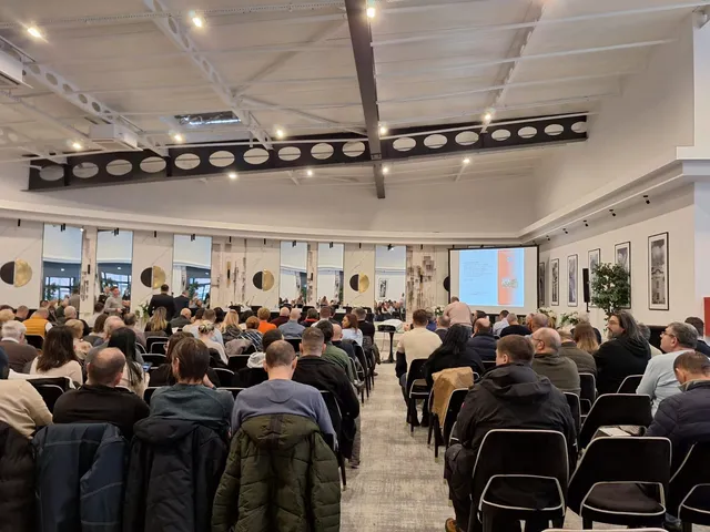 O mulțime de persoane stă așezată pe scaune într-o sală de conferințe cu tavan înalt și un ecran de proiecție luminos în fundal.