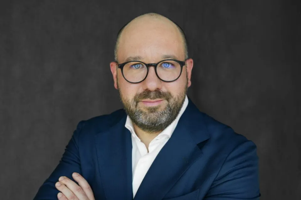 Un bărbat cu ochelari și barbă poartă un sacou albastru peste o cămașă albă, stând cu brațele încrucișate în fața unui fundal gri închis.
