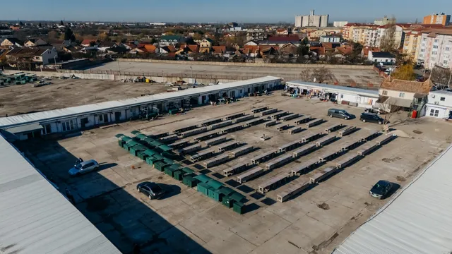 O vedere aeriană prezintă un teren plat cu mai multe containere verzi și negre dispuse în rânduri, lângă câteva clădiri joase și mașini parcate.