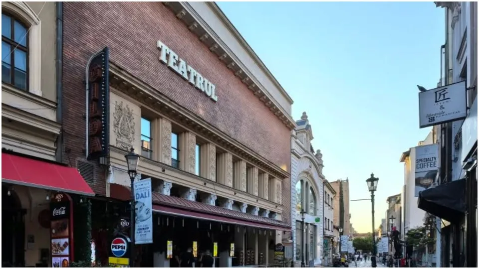 O clădire din cărămidă cu inscripția TEATRUL este situată pe o stradă cu trotuare, fiind încadrată de alte construcții și felinare.