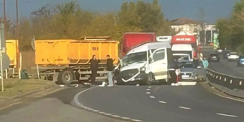 Un microbuz alb și un camion galben sunt oprite pe marginea unui drum asfaltat lângă un vehicul de transport de culoare roșie.