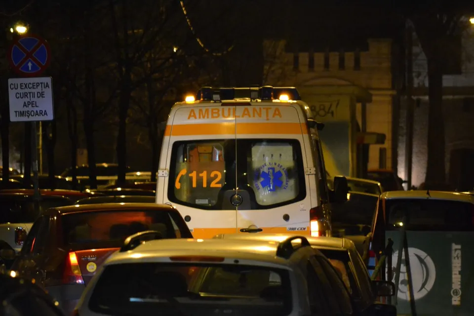 O ambulanță albă cu inscripția AMBULANȚA și numărul 112 este parcată pe un drum noapte, înconjurată de mai multe autoturisme.