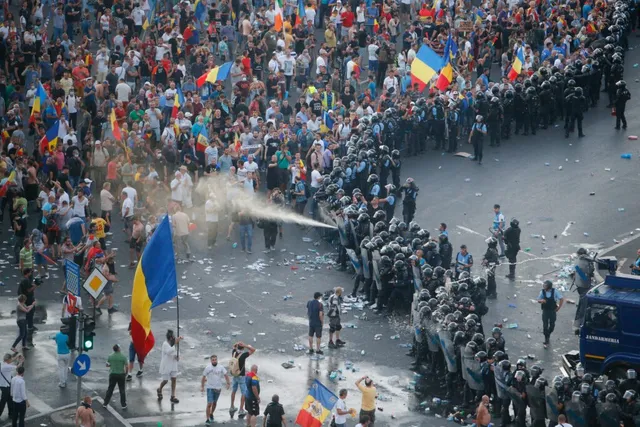 O mulțime de oameni stă în fața unui cordon de polițiști în uniformă în mijlocul unei străzi, în timp ce un jet de apă este proiectat spre mulțime.