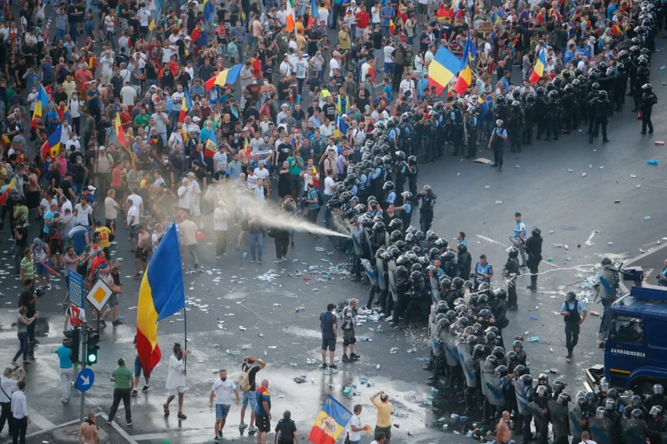 O mulțime de oameni stă în fața unui cordon de polițiști în uniformă în mijlocul unei străzi, în timp ce un jet de apă este proiectat spre mulțime.