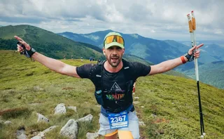 Un bărbat cu brațele întinse poartă un rucsac negru, o șapă cu ochelari și un număr de concurs pe pantaloni în timp ce ține bețe de trekking.
