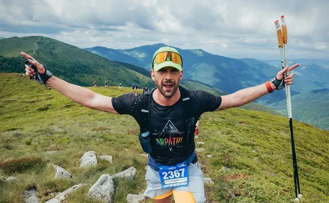 Un bărbat cu brațele întinse poartă un rucsac negru, o șapă cu ochelari și un număr de concurs pe pantaloni în timp ce ține bețe de trekking.