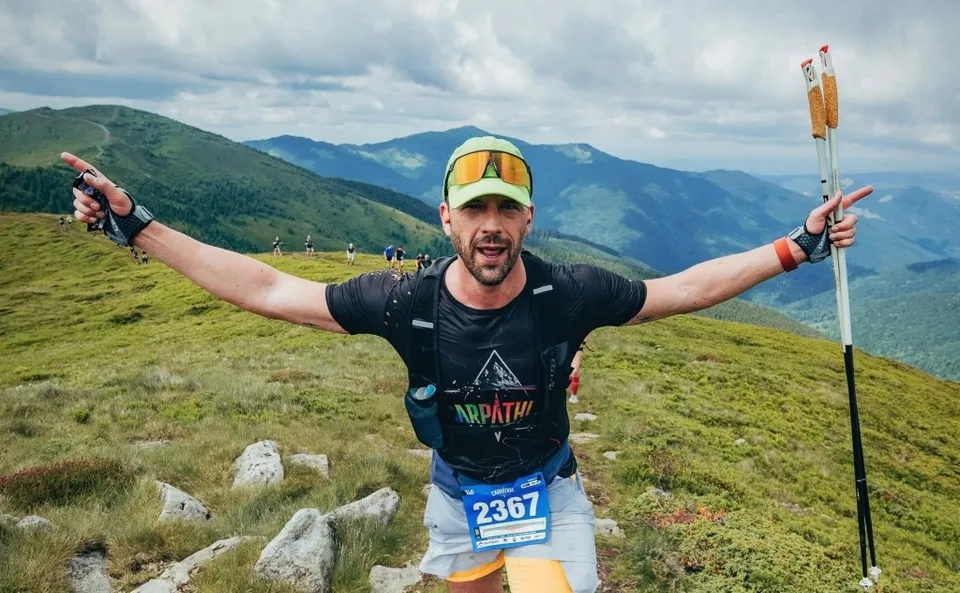 Un bărbat cu brațele întinse poartă un rucsac negru, o șapă cu ochelari și un număr de concurs pe pantaloni în timp ce ține bețe de trekking.