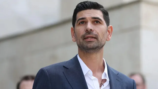 Un bărbat cu părul scurt și barbă stufoasă poartă un sacou închis la culoare peste o cămașă albă deschisă la guler.