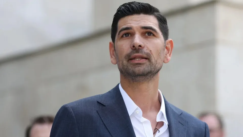 Un bărbat cu părul scurt și barbă stufoasă poartă un sacou închis la culoare peste o cămașă albă deschisă la guler.