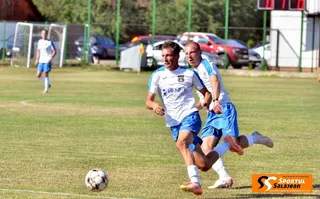 Doi jucători de fotbal în echipamente albastre și albe aleagă pe un teren de iarbă lângă un balon.