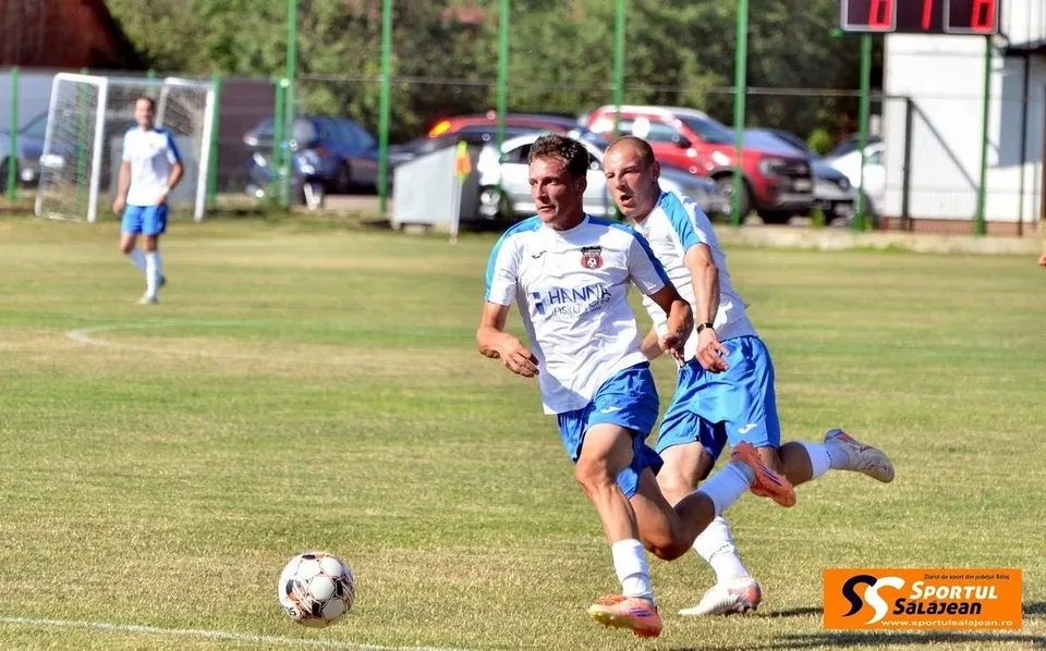Un jucător de fotbal în echipament alb și albastru conduce mingea pe un teren de iarbă, fiind urmărit de un alt jucător în echipament similar.