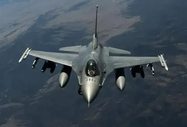 Un avion de luptă gri zboară deasupra unui peisaj de terenuri întinse sub un cer înnorat.