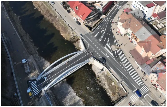 O vedere aeriană prezintă un pod cu arcuri metalice peste un râu, conectând două zone urbane cu străzi asfaltate și clădiri cu acoperișuri colorate.