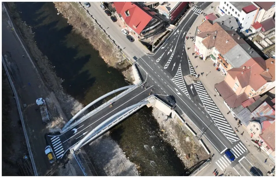 O vedere aeriană prezintă un pod cu arcuri metalice peste un râu, conectând două zone urbane cu străzi asfaltate și clădiri cu acoperișuri colorate.