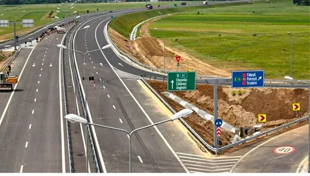 O autostradă cu mai multe benzi de circulație se întinde printre câmpuri verzi, având indicatoare rutiere și stâlpi de iluminat pe marginea drumului.