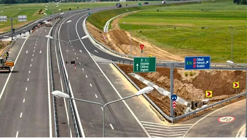 O autostradă cu mai multe benzi de circulație se întinde printre câmpuri verzi, având indicatoare rutiere și stâlpi de iluminat pe marginea drumului.