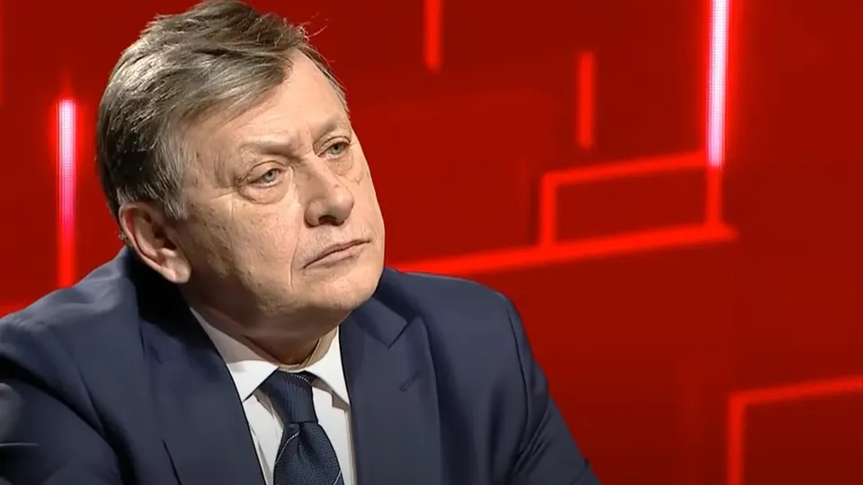 Un bărbat cu părul cărunt, purtând sacou albastru, cămașă albă și cravată închisă, privește spre lateral pe un fundal roșu iluminat.