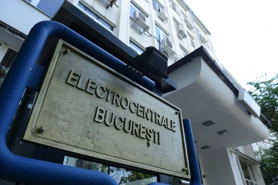 Un semn metalic cu textul ELECTROCENTRALE BUCUREȘTI este montat pe un cadru albastru în fața unei clădiri albe cu unități de aer condiționat.