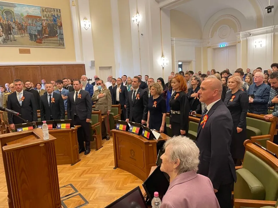 Un grup de persoane îmbrăcate în haine elegante stă în picioare într-o sală de conferințe cu panouri de lemn și scaune verzi.
