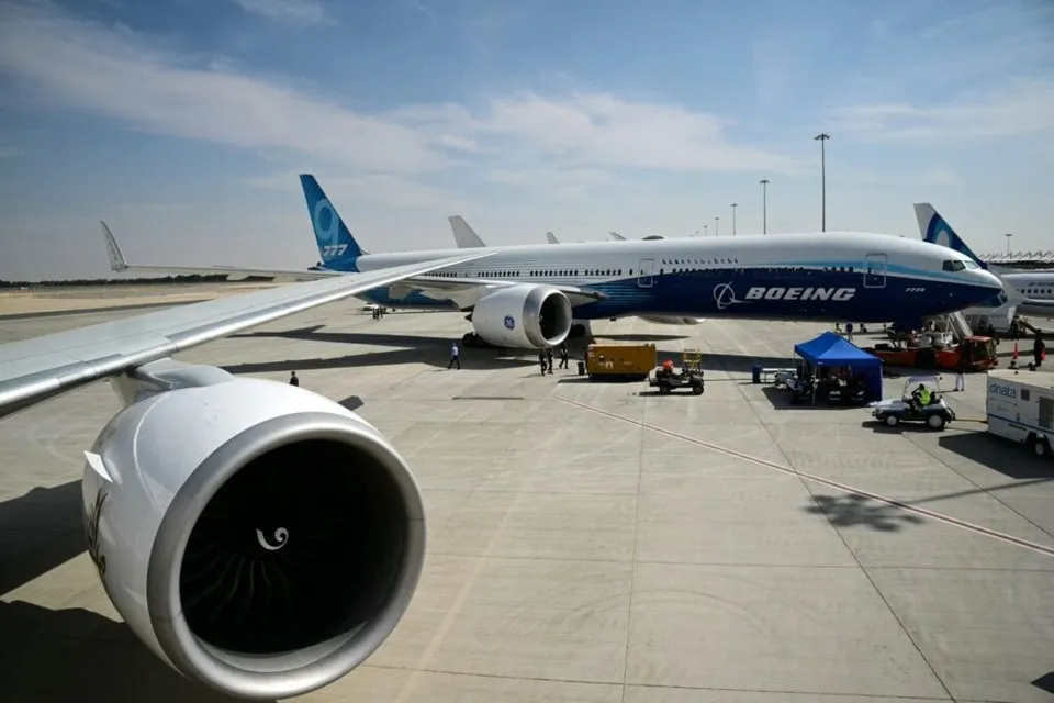 Un avion Boeing alb și albastru stă pe o pistă de aeroport lângă un motor de avion în prim-plan.