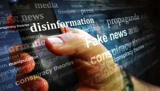 O mână ține un smartphone pe care sunt suprapuse cuvinte precum disinformație, propaganda și fake news pe un fundal întunecat cu diverse texte.