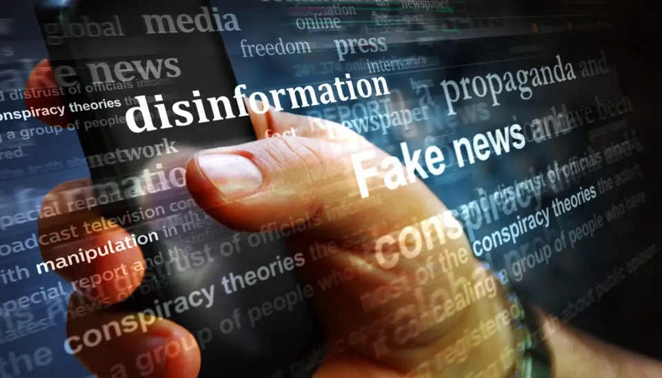 O mână ține un smartphone pe care sunt suprapuse cuvinte precum disinformație, propaganda și fake news pe un fundal întunecat cu diverse texte.
