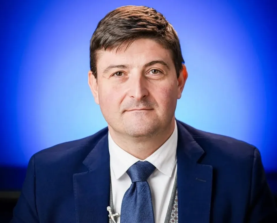 Un bărbat cu părul scurt și brunet poartă sacou albastru, cămașă albă și cravată albastră în fața unui fundal albastru.