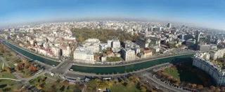 O vedere panoramică de la înălțime asupra unui oraș cu clădiri diverse, un canal și zone cu vegetație sub un cer senin.