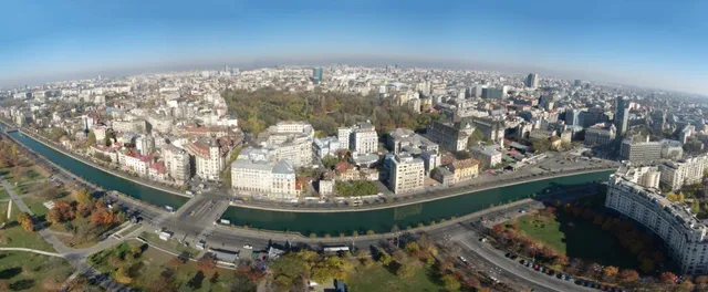 O vedere panoramică de la înălțime asupra unui oraș cu clădiri diverse, un canal și zone cu vegetație sub un cer senin.