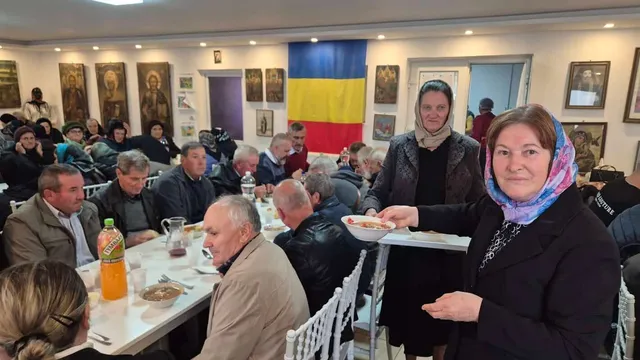 O femeie cu batic într-un cap și o haină neagră oferă un bol cu mâncare unor persoane așezate la mese lungi într-o încăpere.