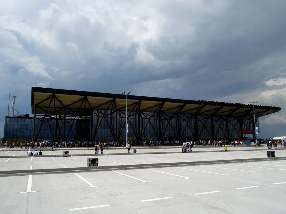 A large modern terminal building stands behind a wide paved area with several small groups of people.