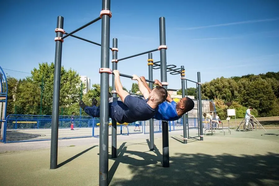 Doi bărbați în tricouri fac exerciții pe barele unui ansamblu de street workout, într-un parc cu copaci și alte aparate în fundal.