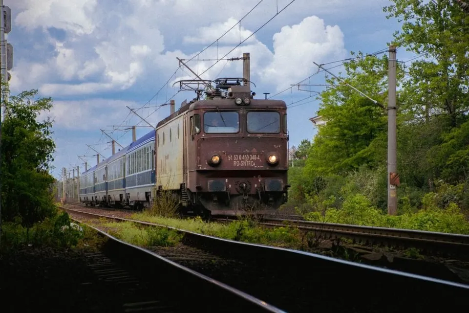 Un tren feroviar cu locomotivă maro și vagoane albastre circulă pe o cale ferată înconjurată de vegetație sub un cer cu nori.