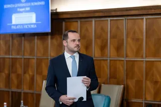 Un bărbat cu barbă scurtă, purtând un costum închis și cravată albastră, ține o foaie de hârtie albă în fața unui panou de lemn.